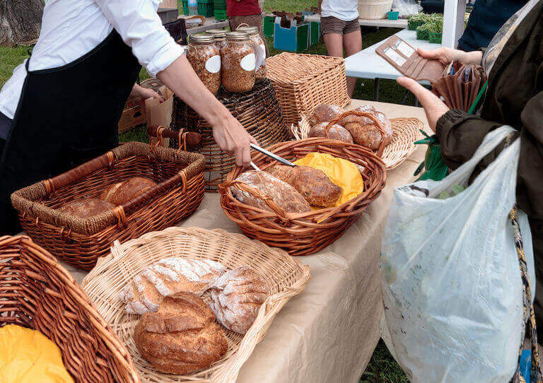 A baker at an outdoor farmers market sells freshly baked organic bread displayed on a table.