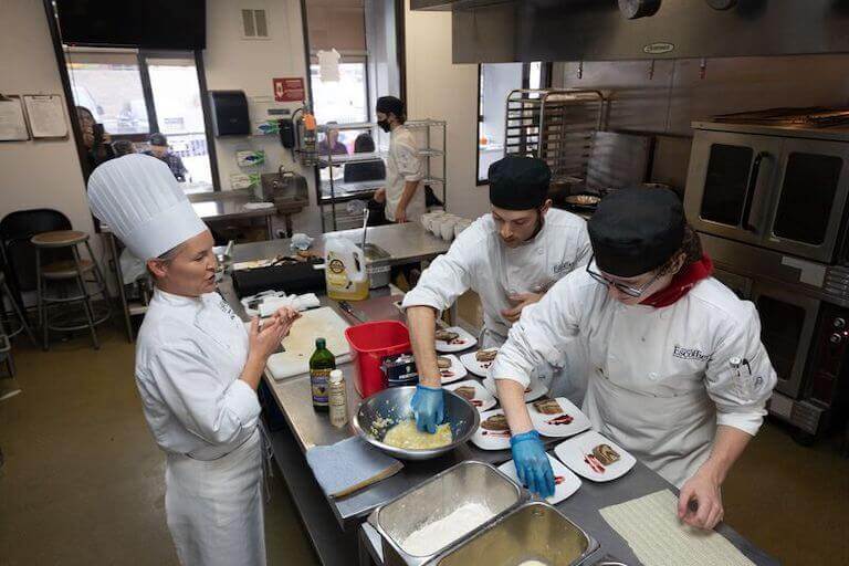 Chef instructor watching two students plate dishes in a kitchen