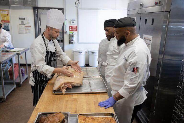 A Chef Instructor shows three students different parts of a cut of meat in a kitchen at Auguste Escoffier School of Culinary Arts Austin.