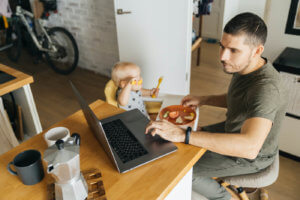 A father feeds his baby while looking at his laptop in the kitchen