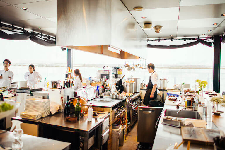 A chef looks over the stove in a large kitchen while other employees work in the distance