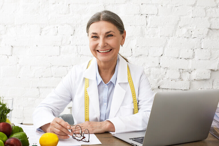 A smiling nutritionist with a measuring tape around her neck works at her desk with a pen and paper and laptop