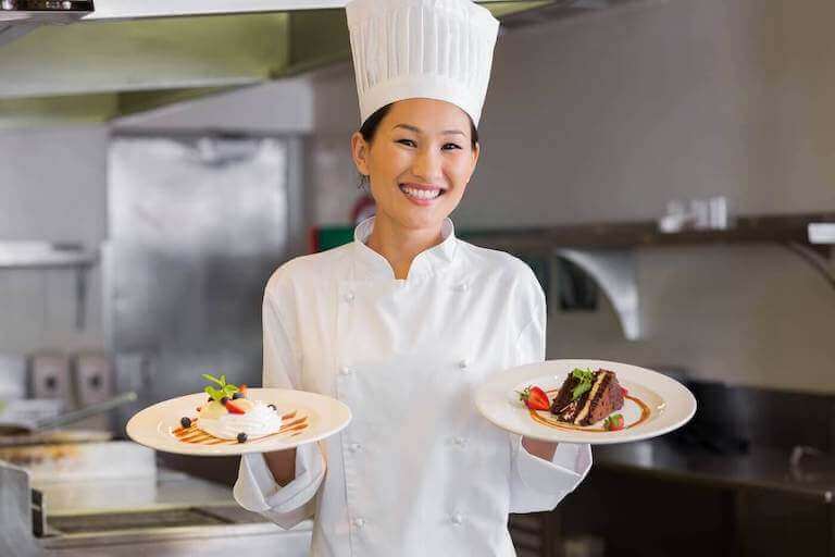 A smiling chef stands in a restaurant kitchen holding two finely plated dessert dishes.