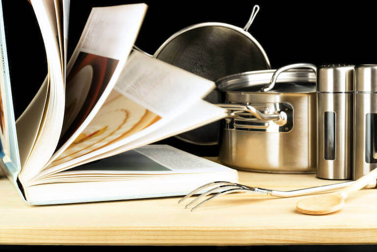 Cookbook, book with pages open next to metal pots and kitchen utensils