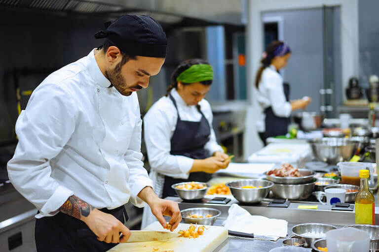 Several chefs work alongside one other chopping and peeling ingredients in a restaurant kitchen.