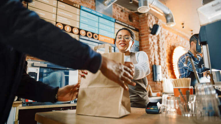 A barista hands a patron a to-go order in a brown paper bag.