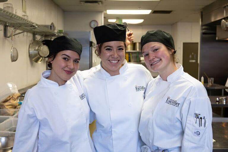 Three Escoffier students in uniform smiling for a photo in a commercial kitchen