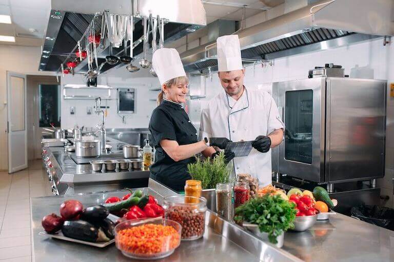 Two chefs standing in a kitchen behind a table full of vegetables discussing the menu