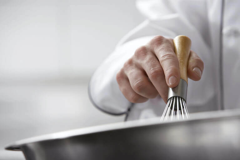 Close up image of a chef's hand holding a whisk in a metal bowl