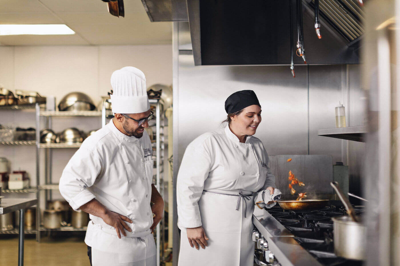 Escoffier student flipping ingredients in a pan while Chef Instructor watches