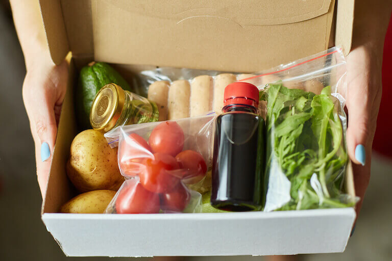 Hands holding a box meal kit with potatoes, tomatoes, herbs, and vinegar