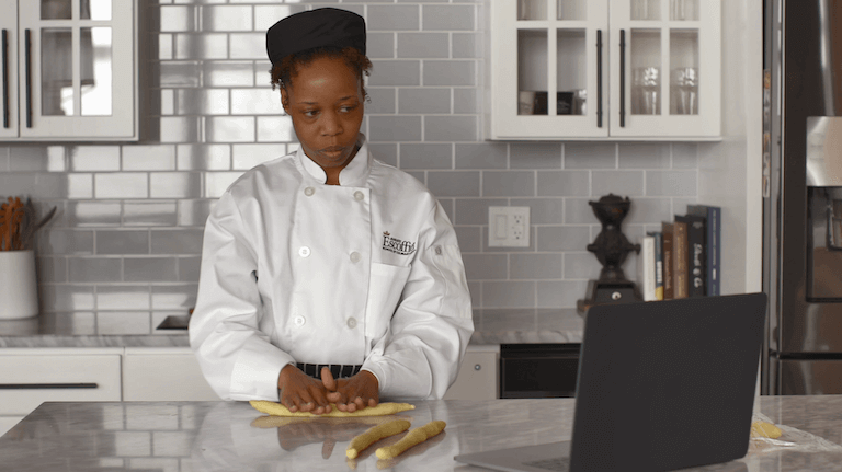 Online student rolling dough in the kitchen while watching computer