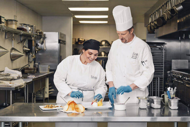 Smiling Escoffier student plates a dish while a chef instructor watches and assists