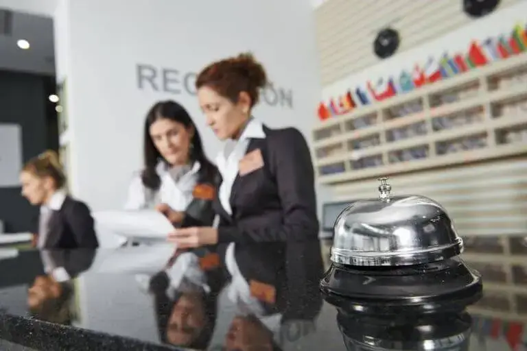 Two workers at a reception desk review notes with a service bell in the foreground.