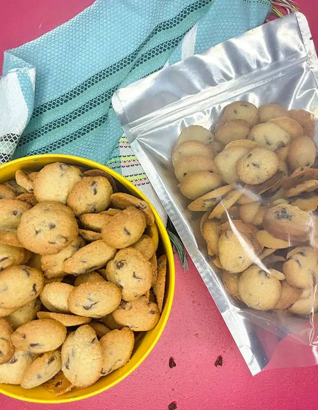 Mini chocolate chip cookies in a yellow bowl and clear bag on a pink table with a teal towel