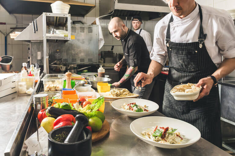 Several chefs work side-by-side plating pasta dishes in a busy restaurant kitchen.