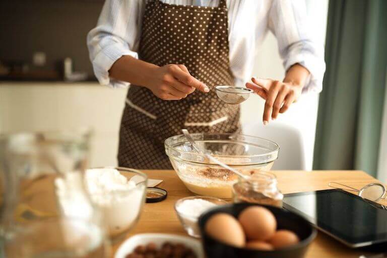 Baker wearing a brown and white apron adding ingredients to a bowl