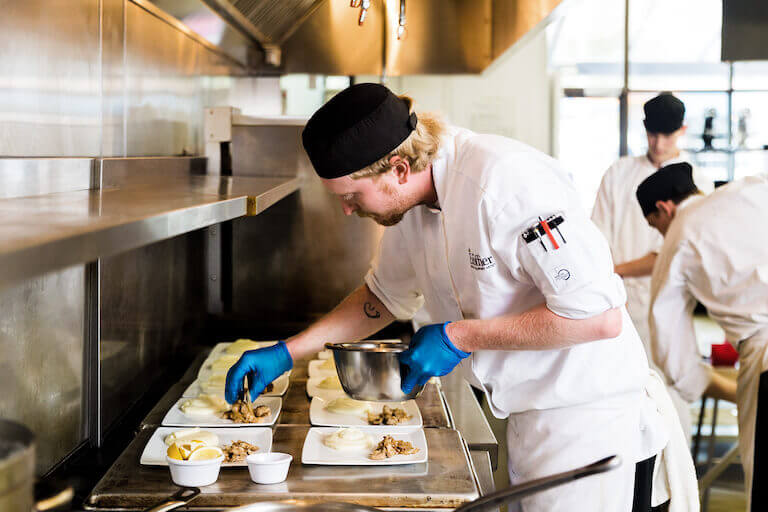 Escoffier student in chef uniform plating meals in a kitchen with other students
