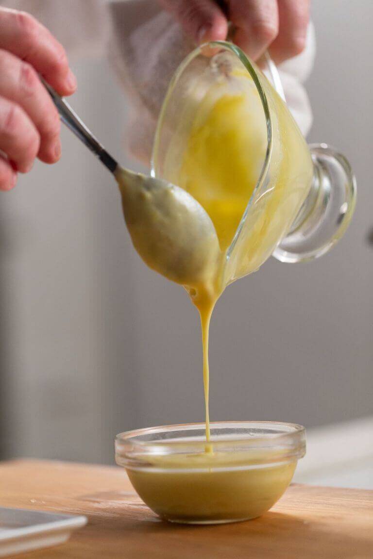 Hollandaise sauce being poured from a glass dish into a small glass bowl