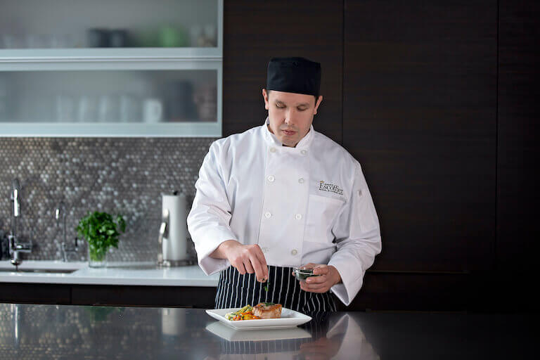 Male culinary student seasoning & garnishing a vegetable and meat dish in home kitchen