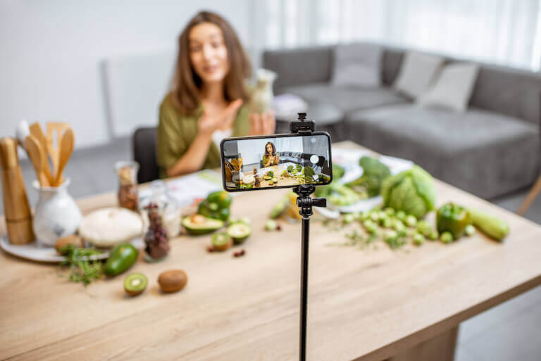 A female food influencer holding a green juice is filmed by a smartphone on a thin stand.