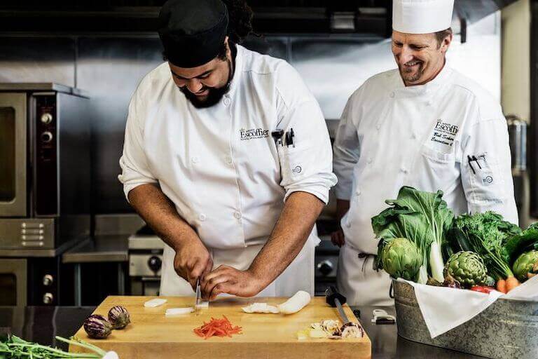Culinary student cutting vegetables while smiling chef instructor observes