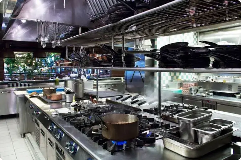 Empty professional kitchen with stainless steel equipment, gas burners, and hanging cookware.
