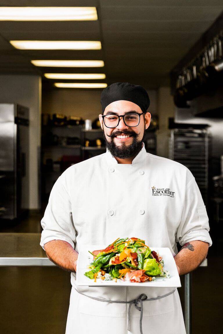 A smiling Escoffier student stands in a training kitchen holding a plated salad