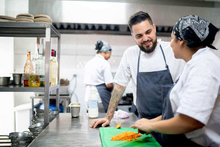 Female cook dicing carrots while male chef with tattoos watches