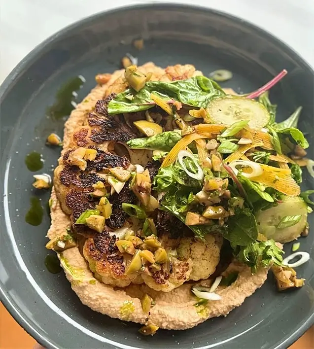 Roasted cauliflower steak on creamy hummus, topped with fresh greens, shaved vegetables, and chopped olives