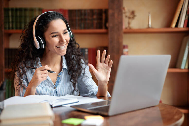 Woman with headphones sitting at a desk smiles and waves to the webcam on her laptop.