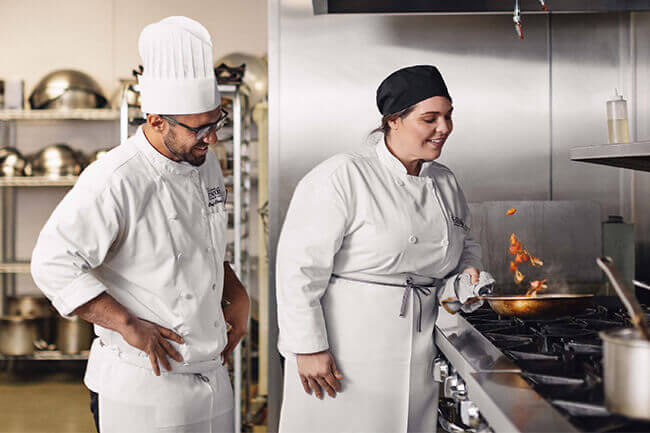 a chef instructor supervising a culinary student over a flaming stove