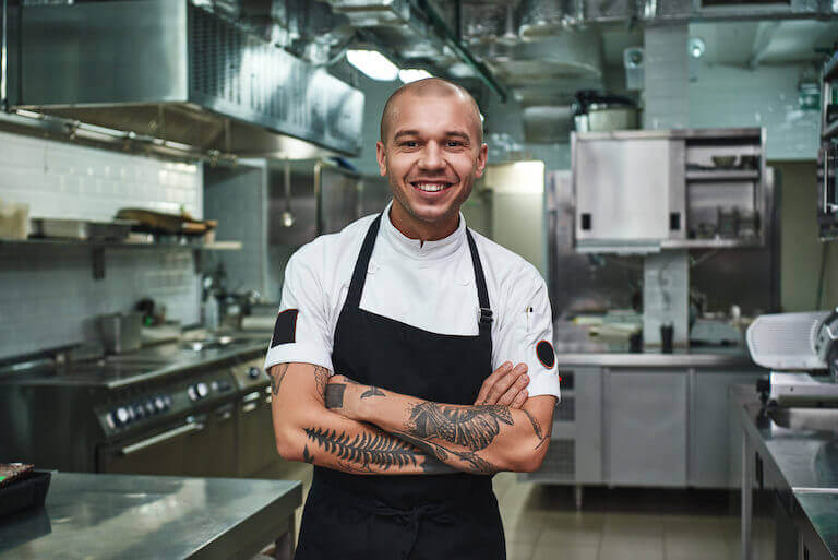 young chef in apron keeping tattooed arms crossed and smiling while standing in a restaurant kitchen