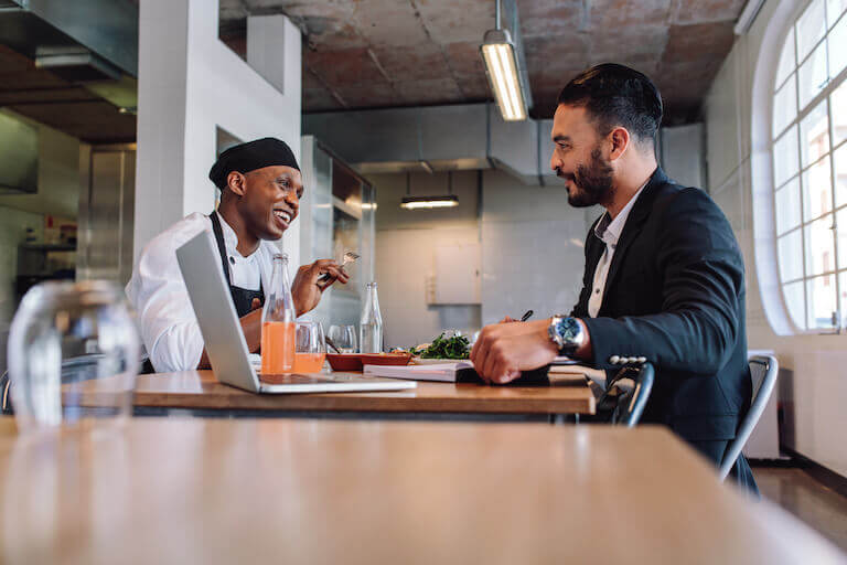 A chef and a restaurant manager sit together at a table in a restaurant dining room, smiling and laughing as the manager makes notes in a notebook.