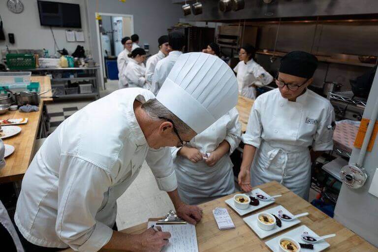 Chef Instructor looks over the work of several students plated on a workbench in a professional kitchen.