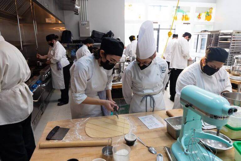 Chef instructor watching as a student cuts dough in a kitchen