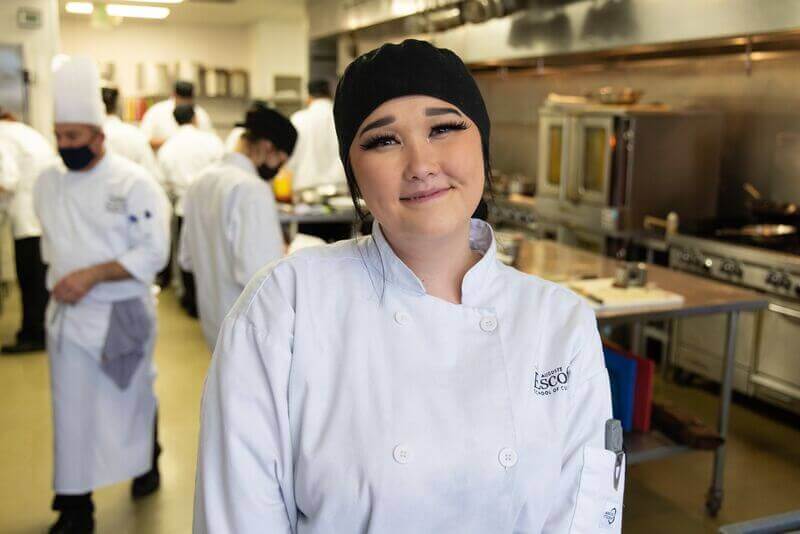 Escoffier student poses for a photo in a kitchen in front of other students cooking