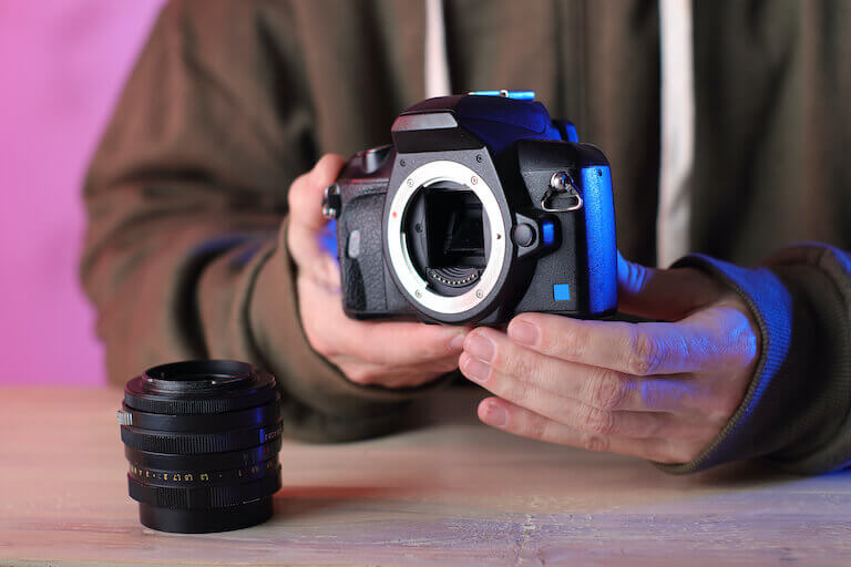 A DSLR in someone’s hands with the lens detached and sitting on the table in front of them. 