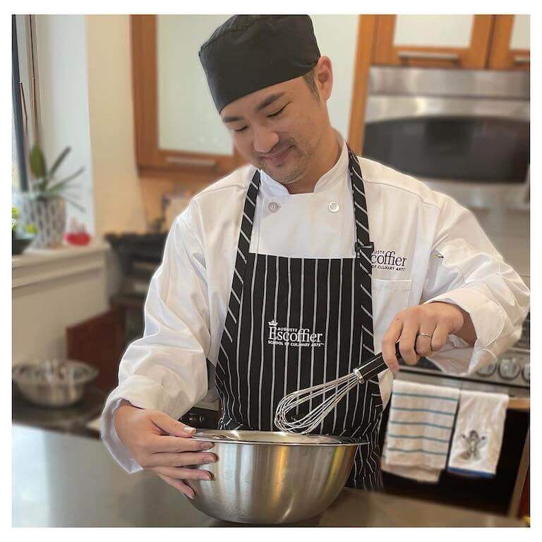 Medium shot of a smiling student in a striped Escoffier apron whisking the contents of a metal bowl.