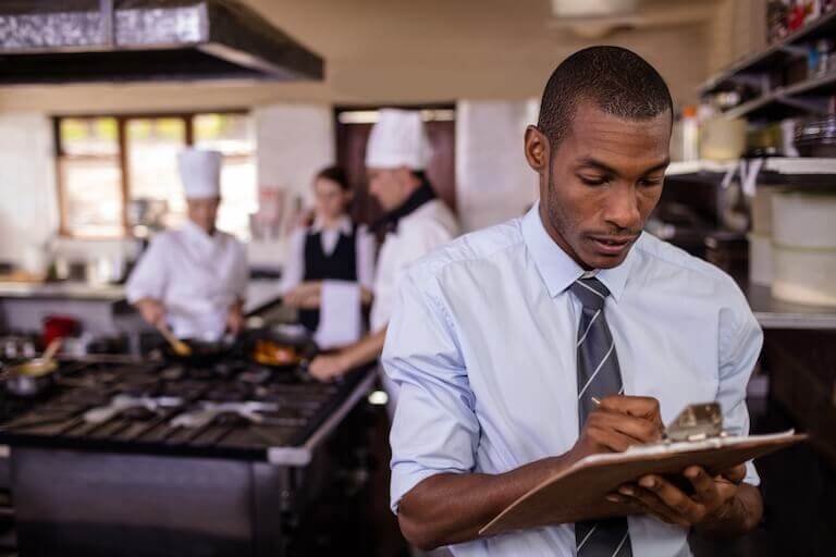 A male restaurant manager in a shirt and tie goes over his business’ accounts while employees work behind him.