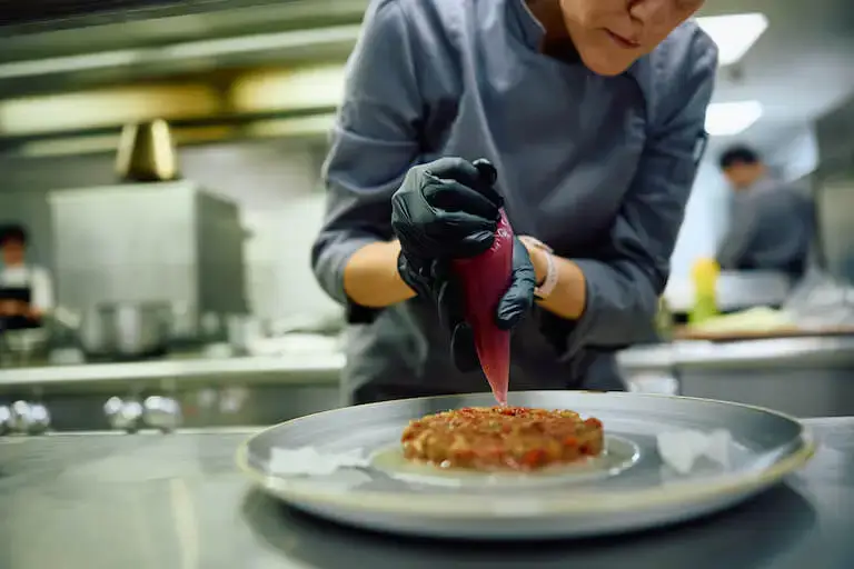 Pastry chef wearing black gloves piping a red sauce or purée onto a plated dessert in a professional kitchen.