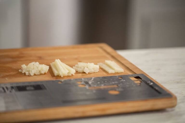 A wooden cutting board on a counter with potatoes that have been cut in four different ways, including julienne.