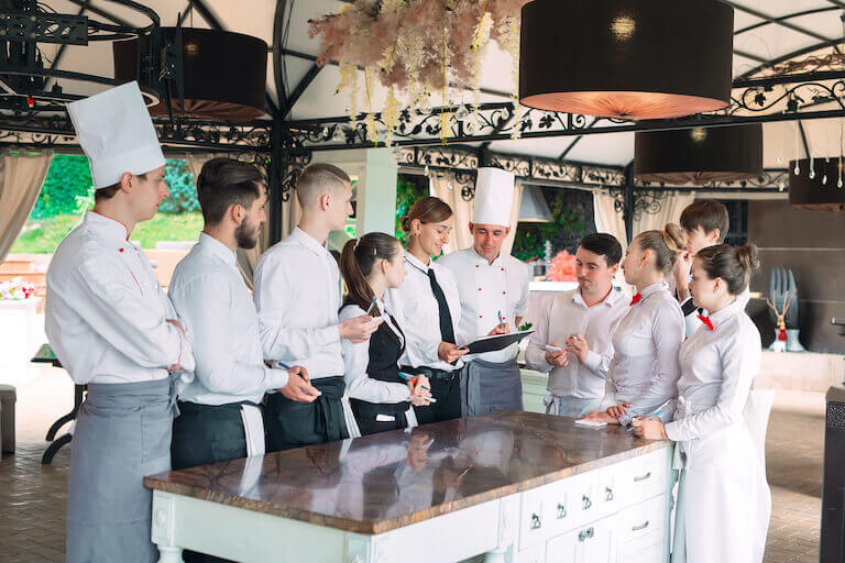 A mix of employees gather in a kitchen including chefs, wait staff, bussers, and hosts.