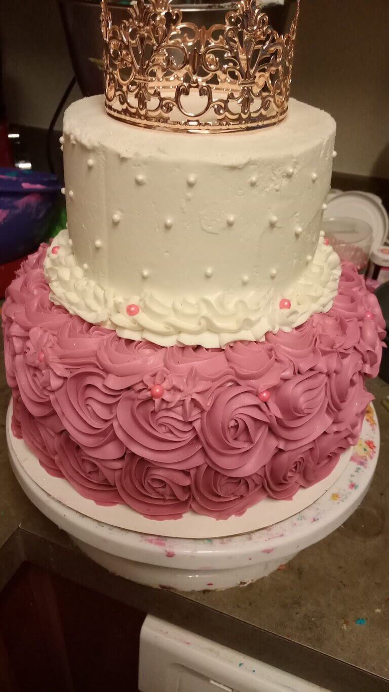 Two tier cake with white frosting and pink flowers topped off with a gold crown