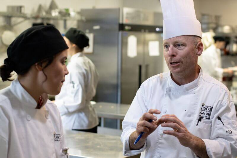 Chef instructor in a white toque and jacket speaks with a culinary student in a professional teaching kitchen, gesturing as stainless steel equipment and other students work in the background.