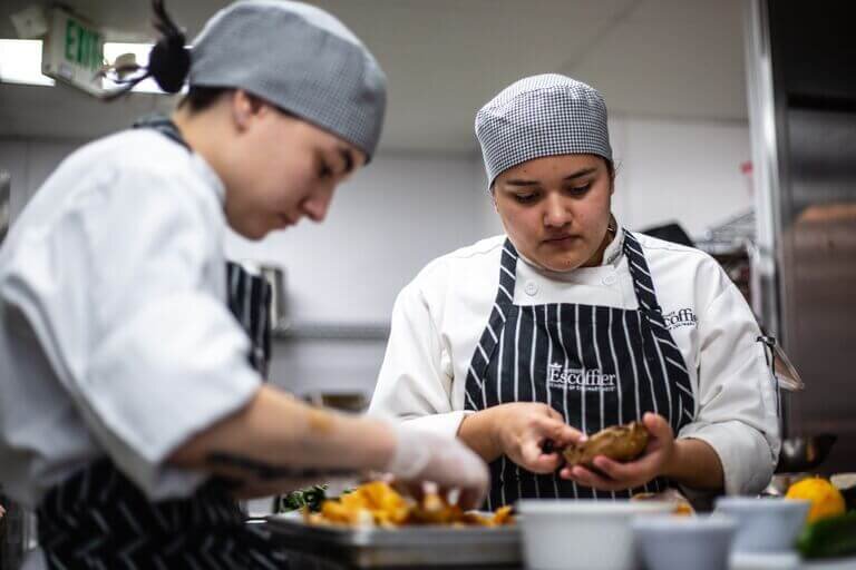 Two culinary students wearing Escoffier-branded aprons and chef’s jackets work with ingredients in a professional kitchen.