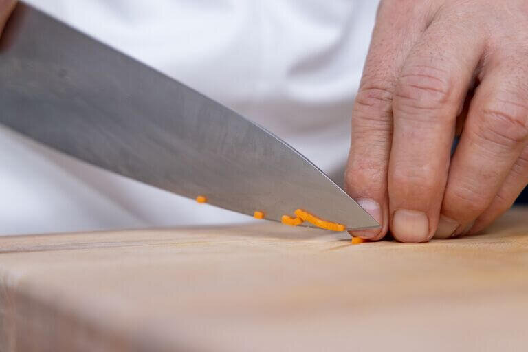 Close-up of a person using a chef's knife on a wooden cutting board to finely dice a carrot into precise brunoise cuts.