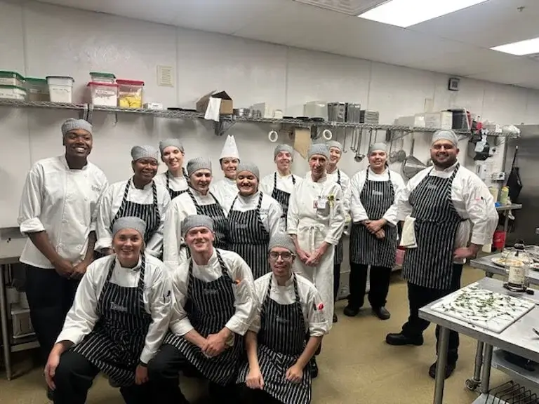 A group of campus ambassadors poses in an on-campus commercial kitchen, most of them wearing the same style of gray houndstooth cap.