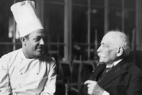 August Escoffier having a conversation with a chef in a traditional chef’s jacket and toque.