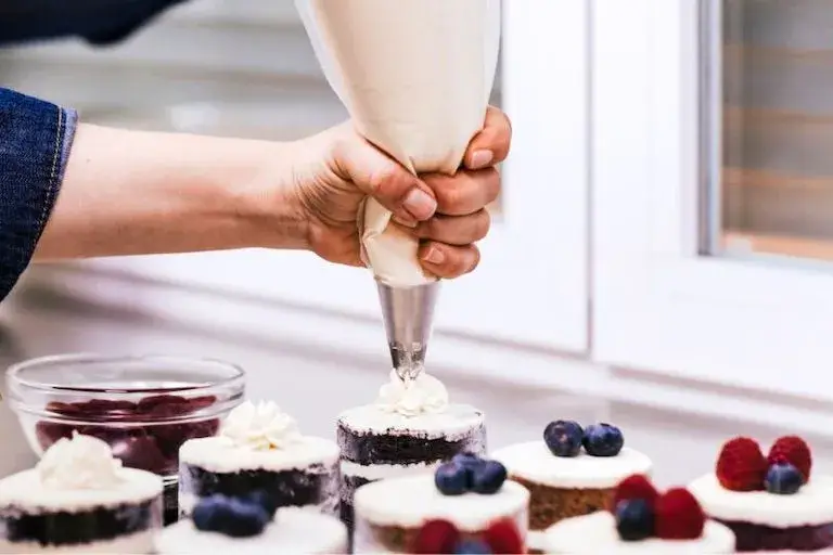 Close-up of a hand piping whipped cream onto mini cakes decorated with berries.